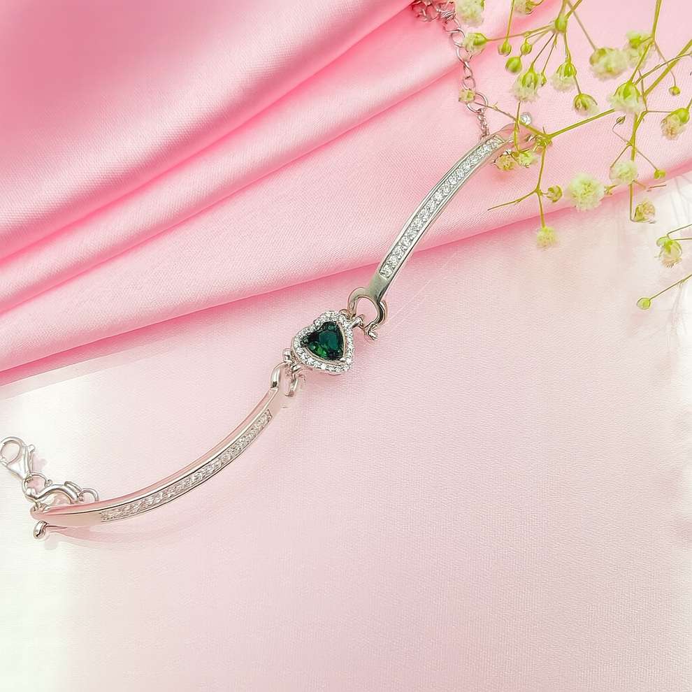 Silver bracelet with a green heart charm on a pink fabric background